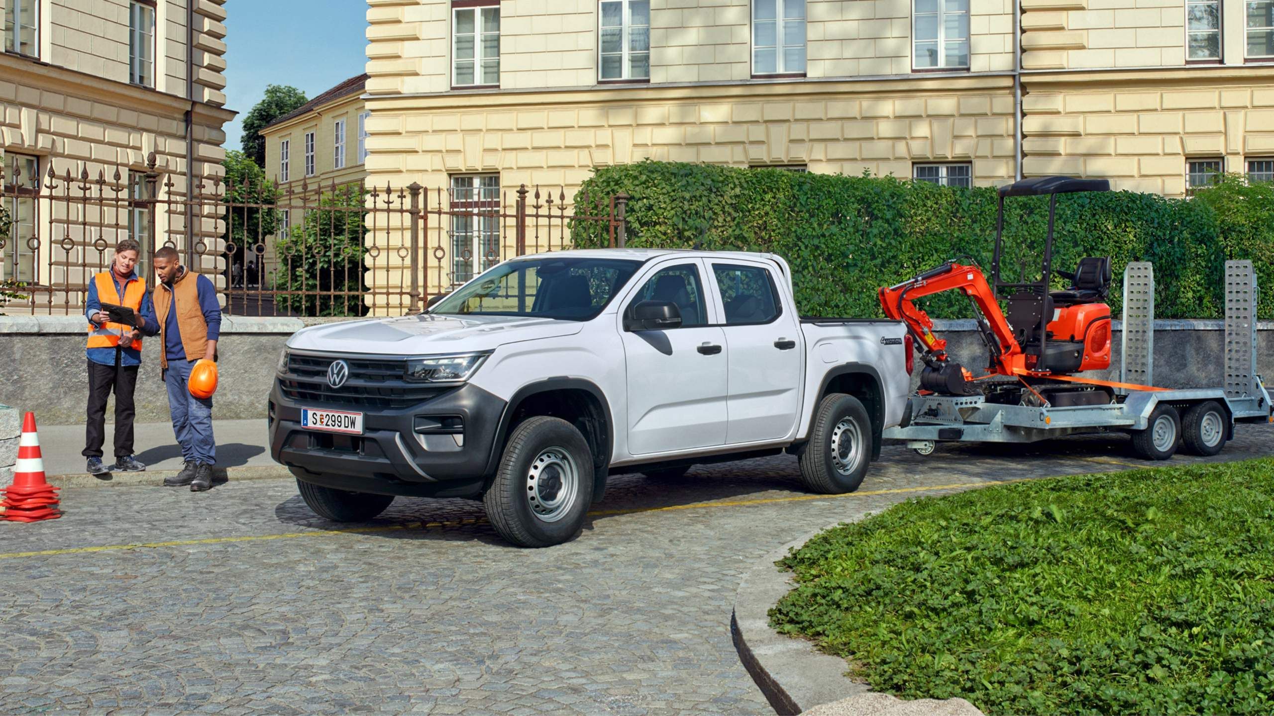 Ein VW Amarok mit Bagger auf dem Anhänger steht auf einer Baustelle und zwei BauarbeiterInnen stehen daneben